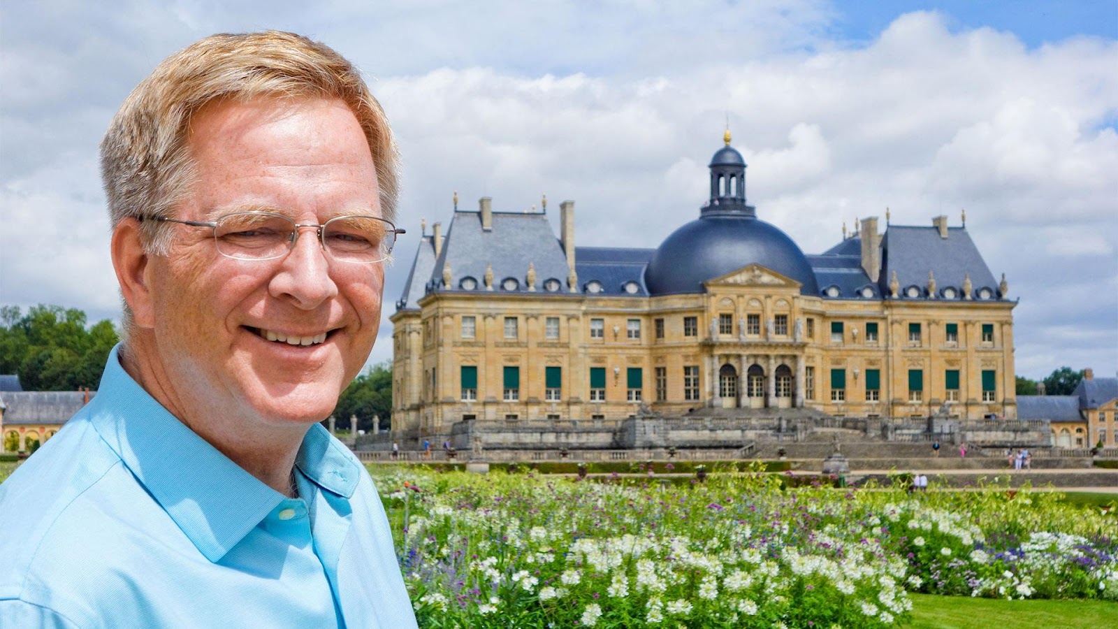 A picture of Rick Steves in front of an old Victorian-style building somewhere in Europe with a beautiful garden in front of it.