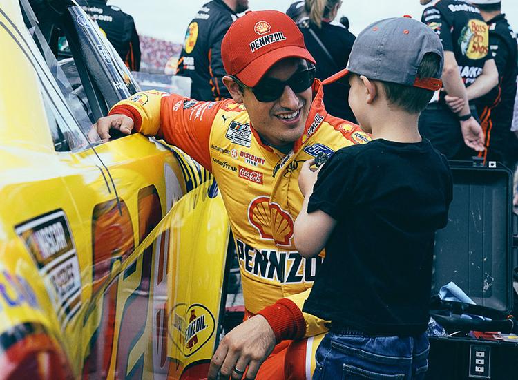 A promotional image of NASCAR driver Joey Logano kneeling next to his racecar with his son Hudson